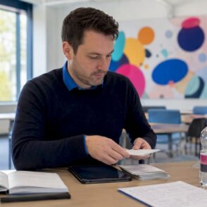 Een IT-trainer zit aan zijn bureau en legt kaartjes en een tablet netjes klaar, zodat alles overzichtelijk is voor de komende les.